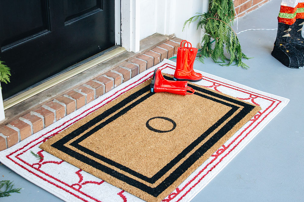 A pair of small red rainboots sits on a monogrammed doormat that is layered on top of a red and white rug.