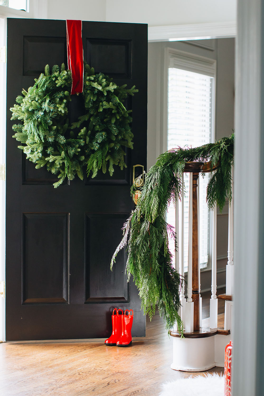 A black front door with a wreath held open by a pair of small red rainboots and a stairway wrapped in garland.