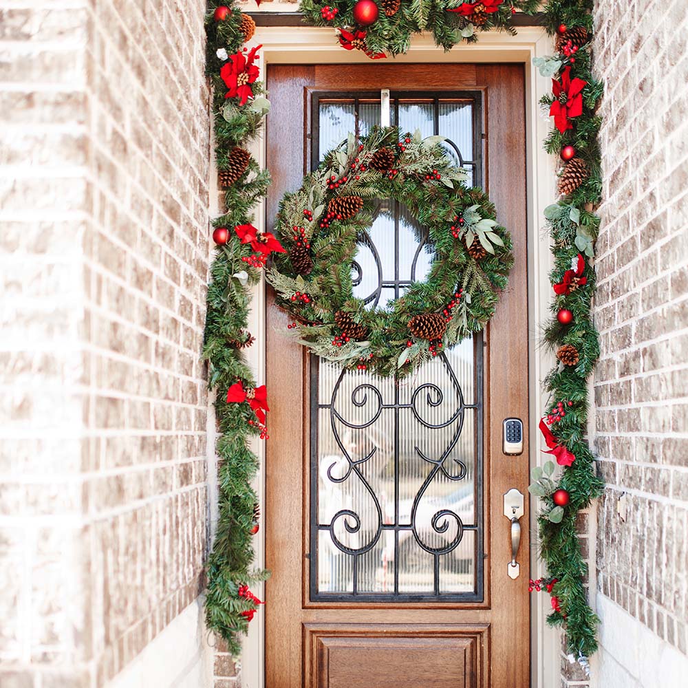 A holiday wreath and garland with red accents decorates a wooden and glass front door.
