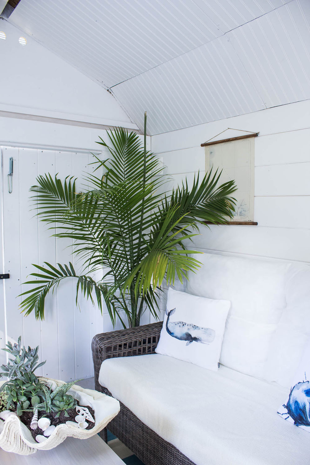 The inside of a "she shed" with white shiplap walls, a couch with decorative pillows, a plant and a table with coastal decor. The inside of a "she shed" with white shiplap walls, a couch with decorative pillows, a plant and a table with coastal decor.
