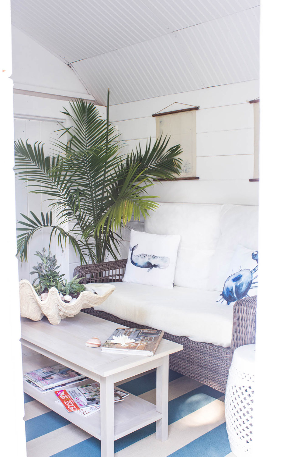 The inside of a "she shed" with white shiplap walls, a wicker couch with decorative pillows, a plant and a table with coastal decor. The inside of a "she shed" with white shiplap walls, a wicker couch with decorative pillows, a plant and a table with coastal decor.