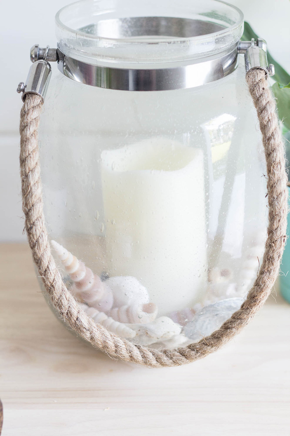 A clear candle holder with a white candle and shells in the bottom sitting on a table. A clear candle holder with a white candle and shells in the bottom sitting on a table.