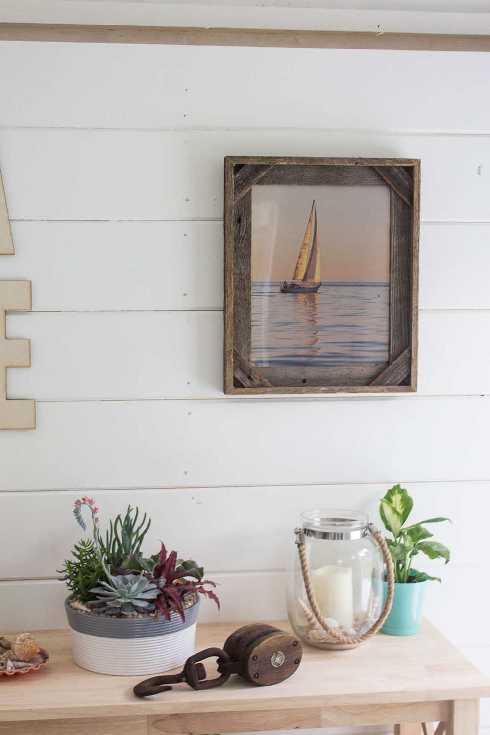 White shiplap wall with a framed photo of a sail boat and a wooden table with plants and a candle. White shiplap wall with a framed photo of a sail boat and a wooden table with plants and a candle.