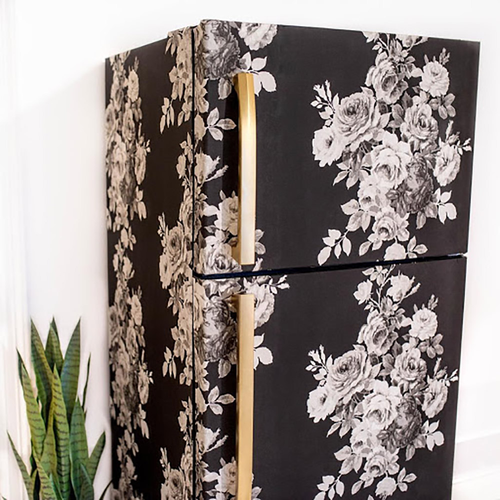 A refrigerator that has been wrapped in a black and floral wallpaper with the door handles sprayed gold.