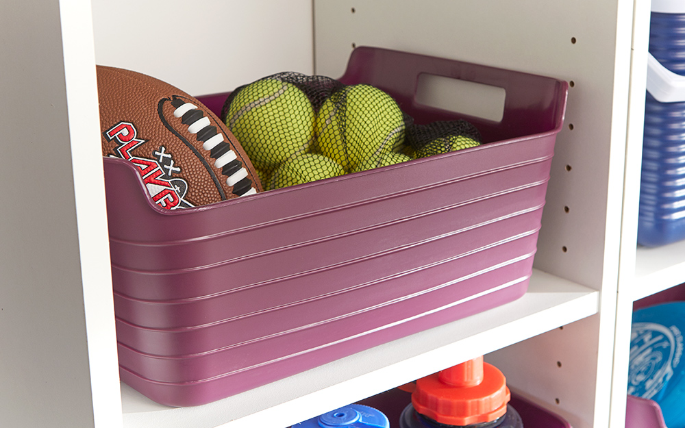 Tennis balls and a football are stored inside a pink bin on a white shelf.