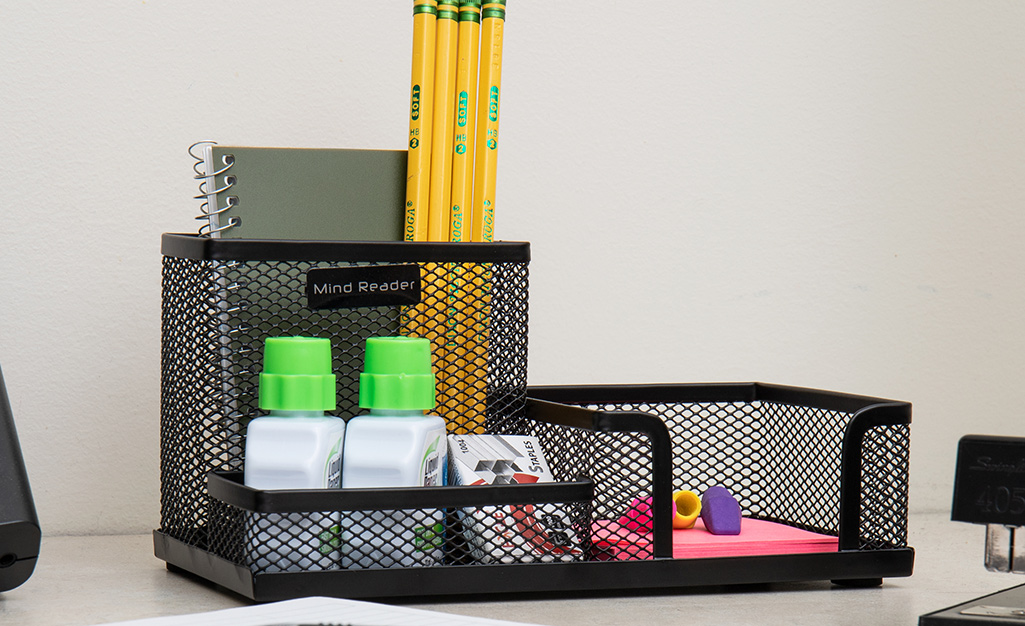 A desk organizer made of wire mesh holds pencils, erasers, a small notebook, sticky notes and other office supplies.