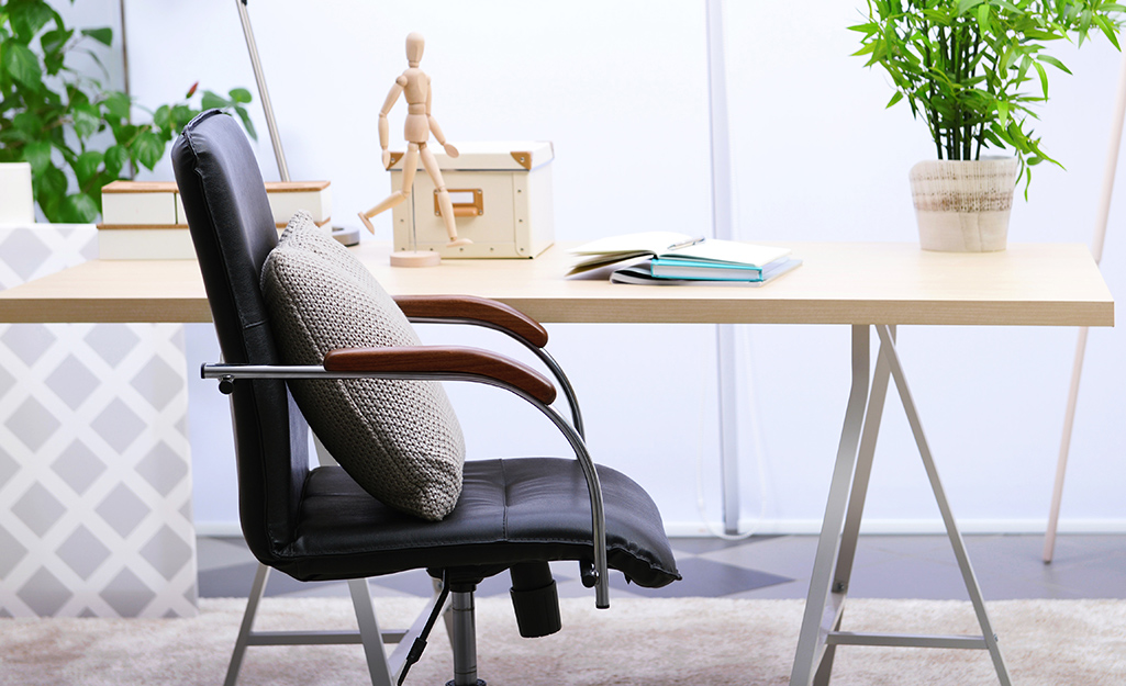A throw pillows leans against the back of an office chair in front of a light wood desk in front of a window.