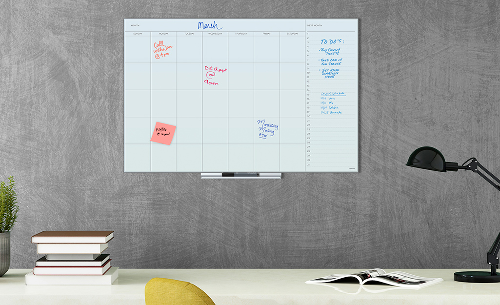 A dry erase calendar hangs above a desk on the wall of a cubicle.