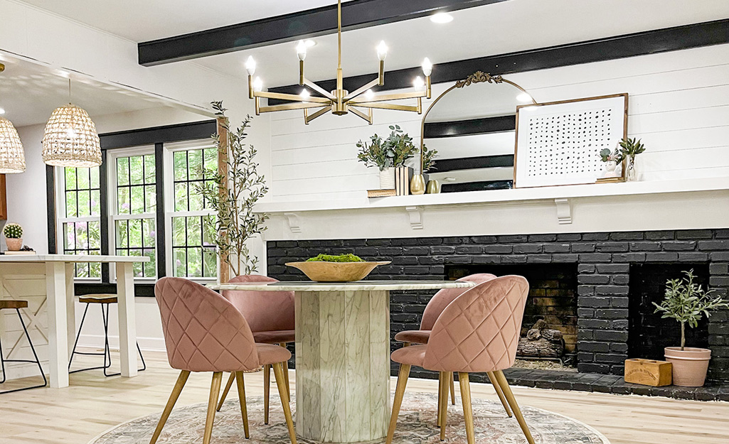 A modern chandelier hangs above a dining table that sits in front of a long fireplace of brick painted black.