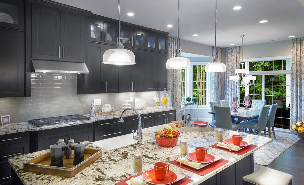 Pendant lights and recessed lighting in the cool tones of daylight bulbs illuminate a kitchen with an island and dark cabinets.