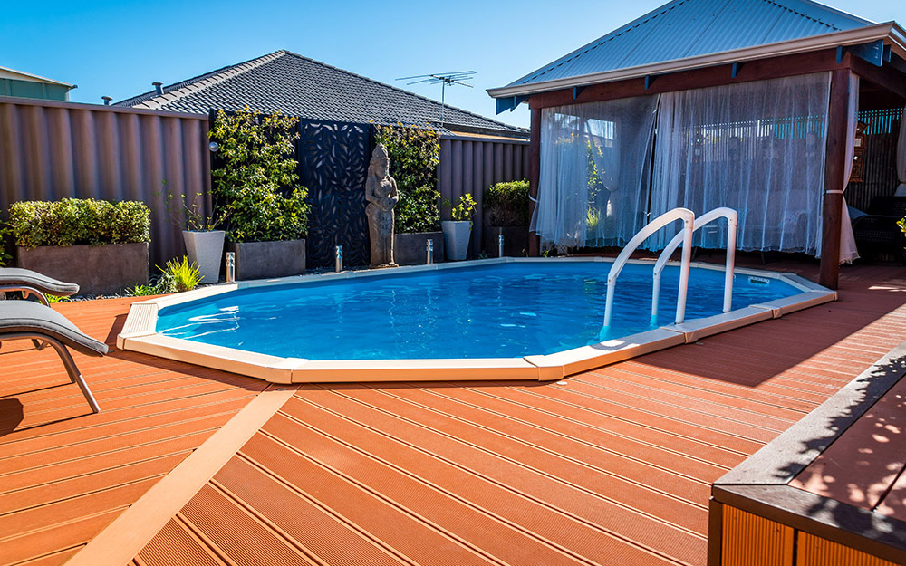 A wooden deck surrounding a swimming pool in the backyard of a brick home.