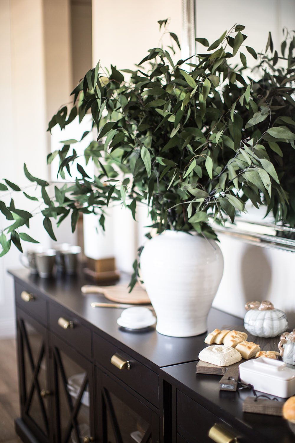 A large white vase with greenery decorates the top of a buffet. A large white vase with greenery decorates the top of a buffet.