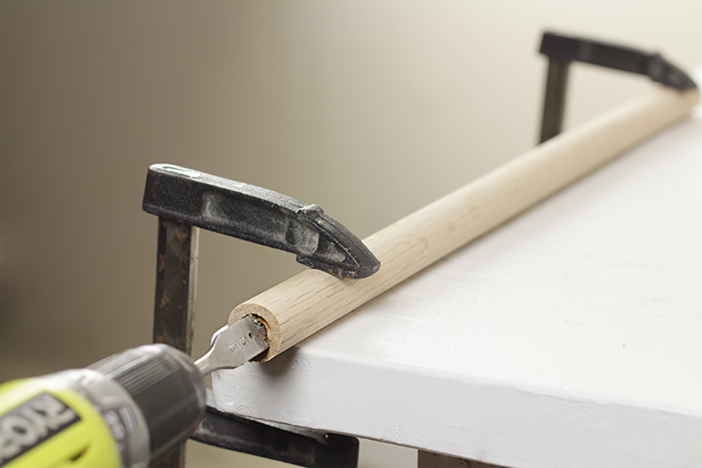 A spade bit drills through a securely clamped dowel on a table. 