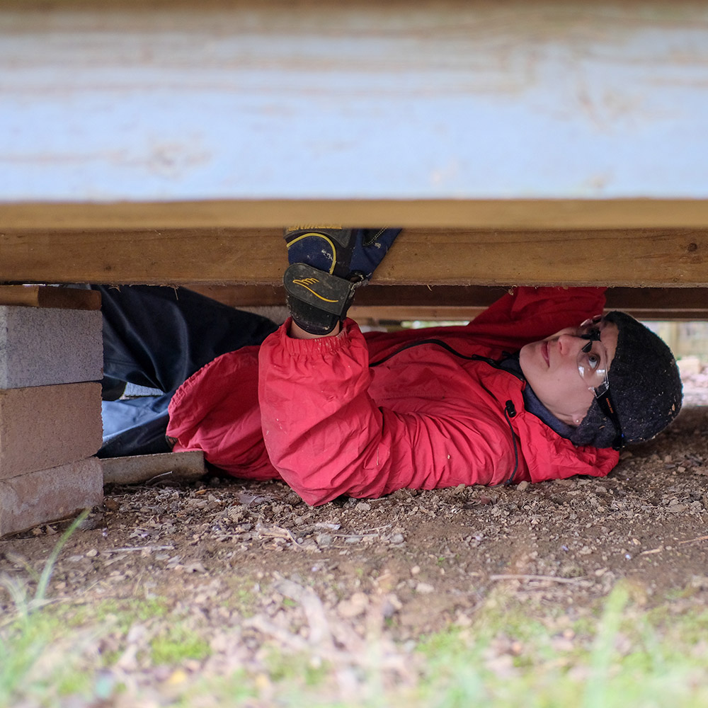 A person in a red jacket working in a crawl space underneath a shed.