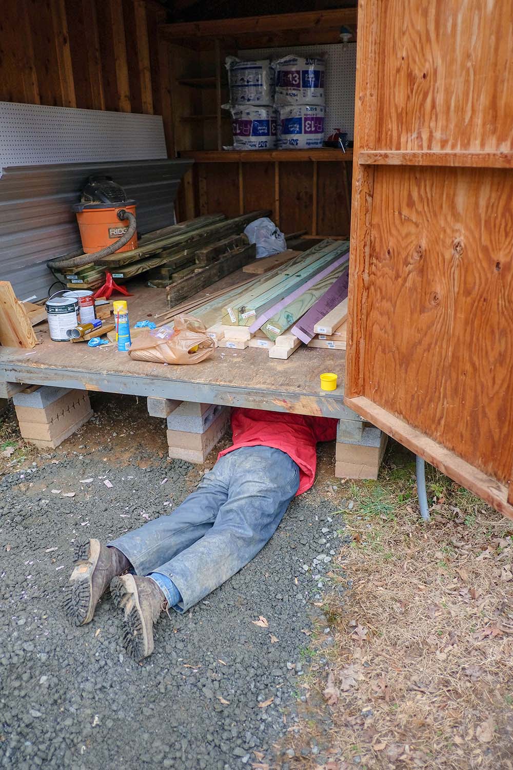 A person's legs hang out from underneath a shed filled with lumber.