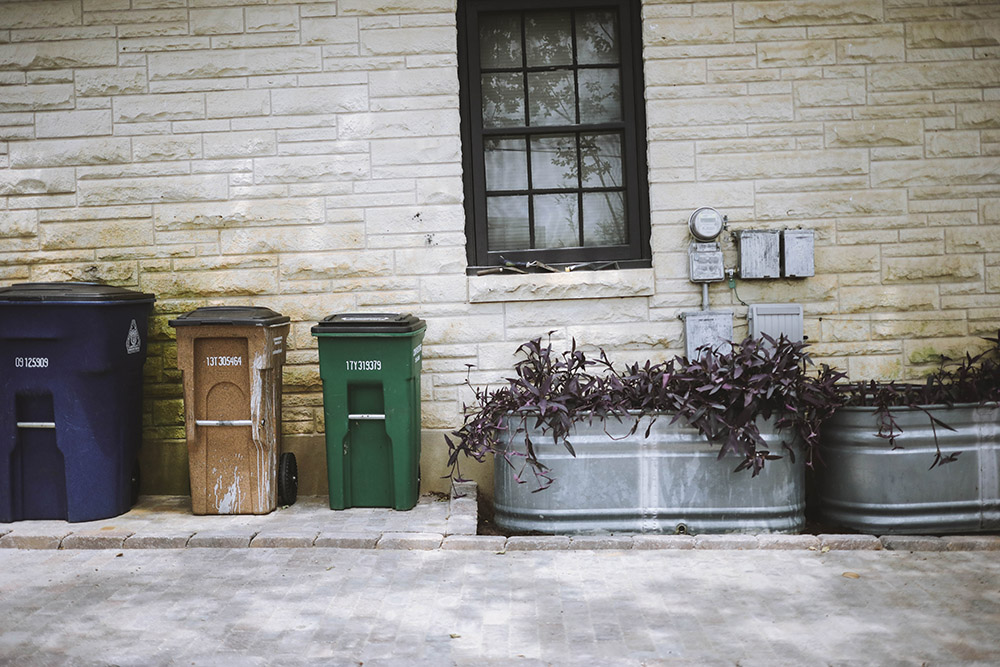 A group of trashcans sit against the wall on the side of a home.