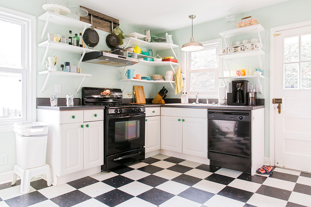 Kitchen with mint green walls, black and white checked flooring, white cabinets, black appliances and white shelves with miscellaneous kitchen items.
