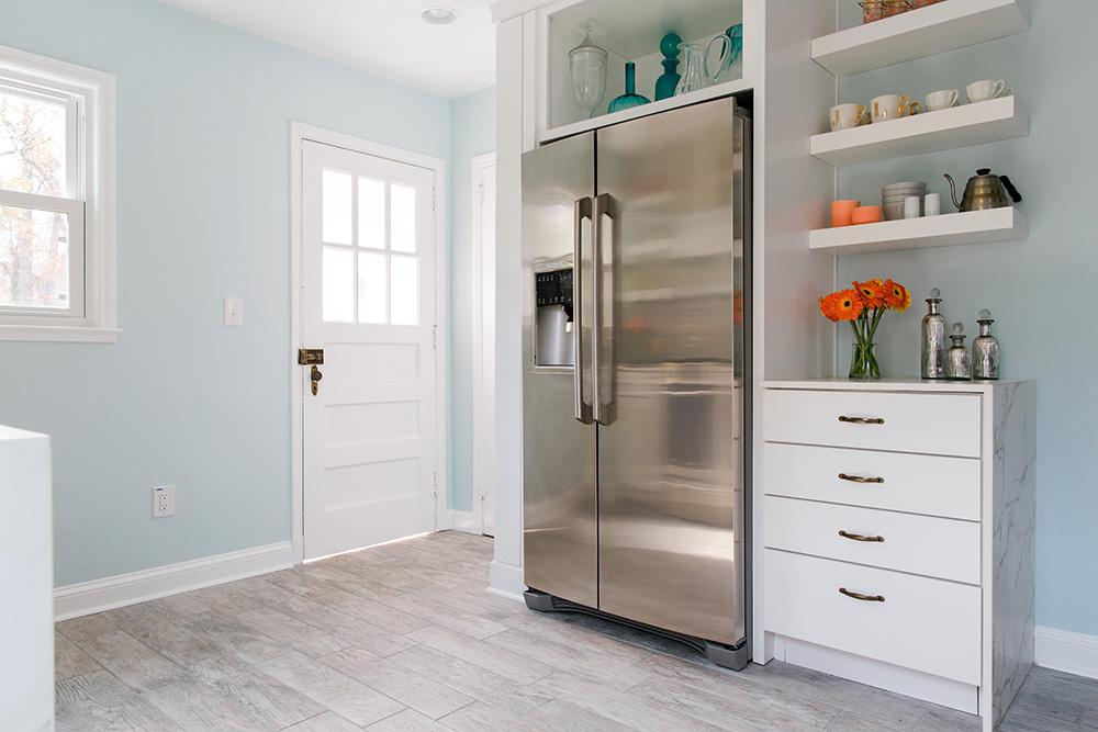 A kitchen with light blue walls, a white door, vinyl flooring, silver refrigerator, white shelving with glassware and white drawers. 