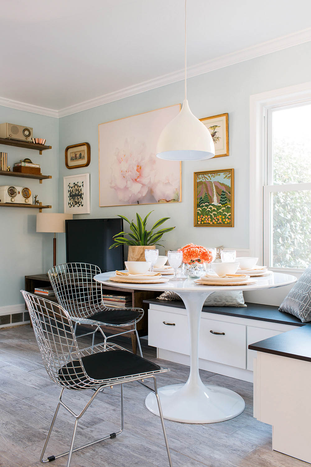 An eating nook with a white table and place settings, two chairs and a bench in a living room space with light blue walls, a television, wall art and lighting.