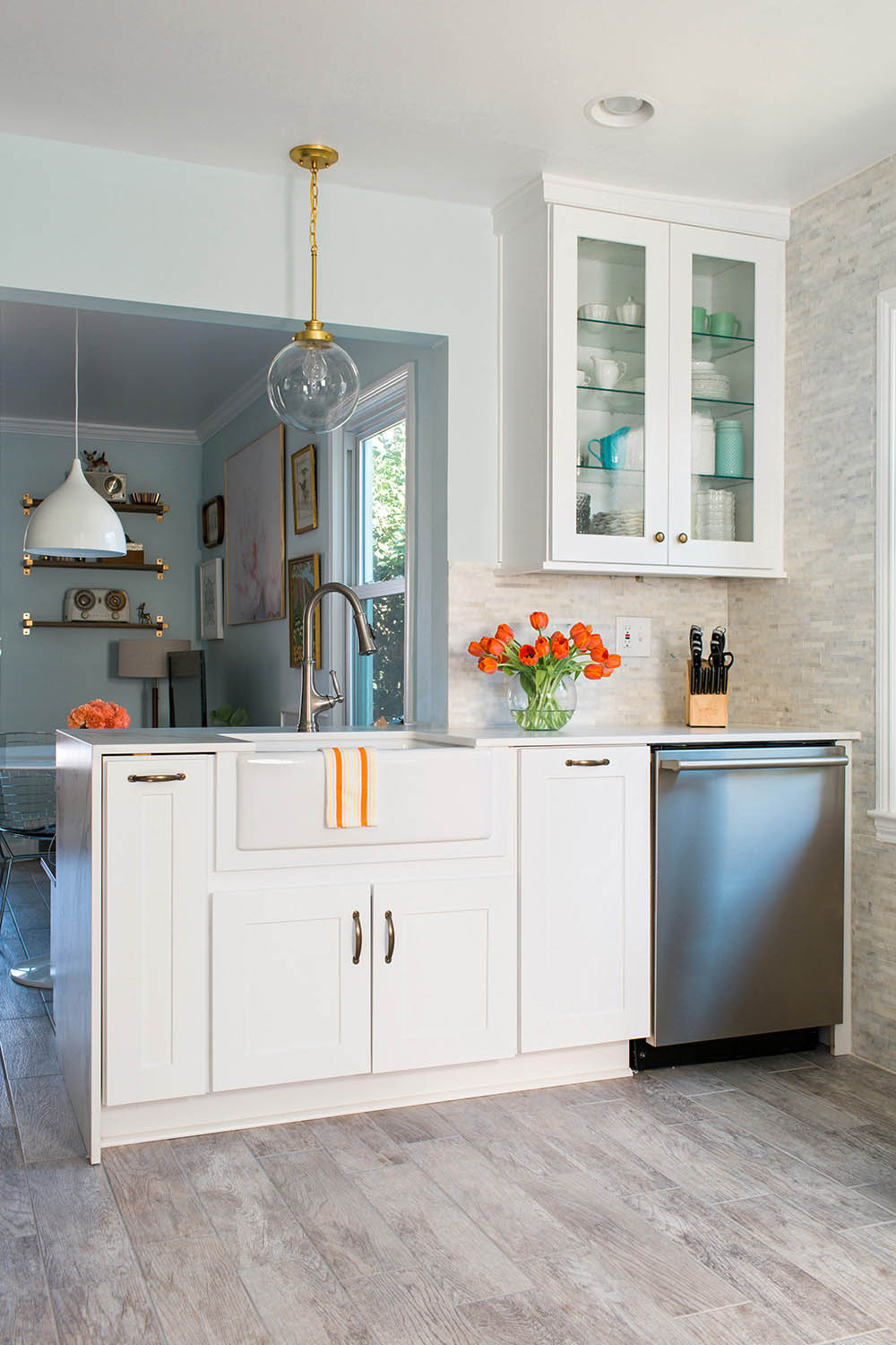 A kitchen with white cabinets and bronze hardware, a hanging light fixture, silver dishwasher and a white cabinet with glassware.