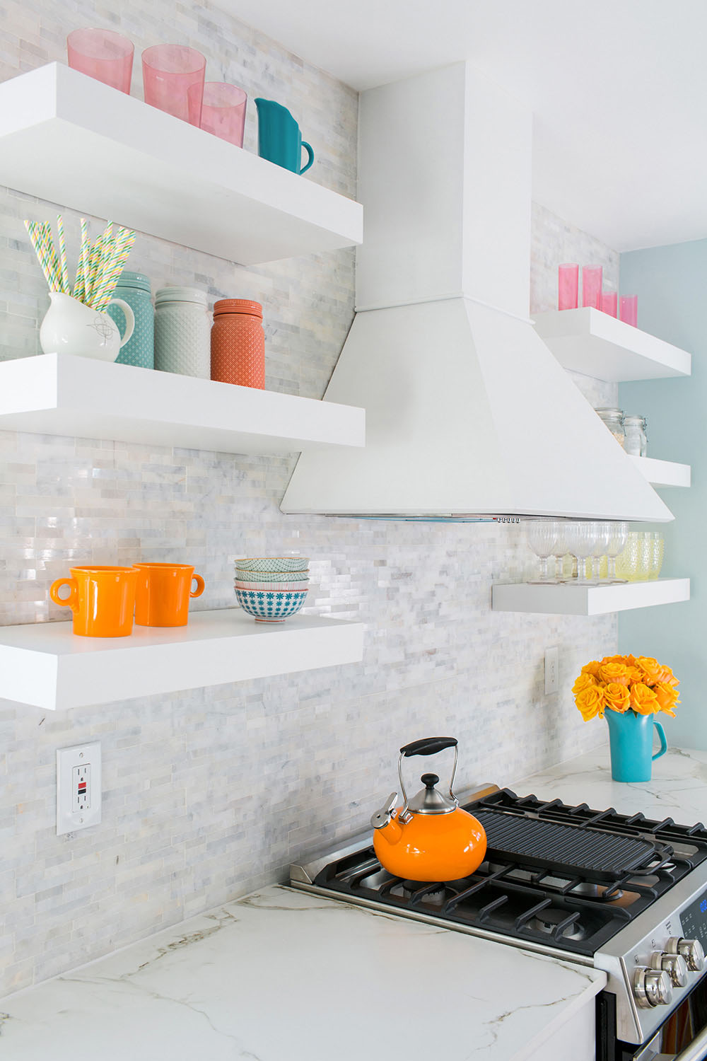A kitchen stove with a white vent hood and white shelving with glassware and vases.