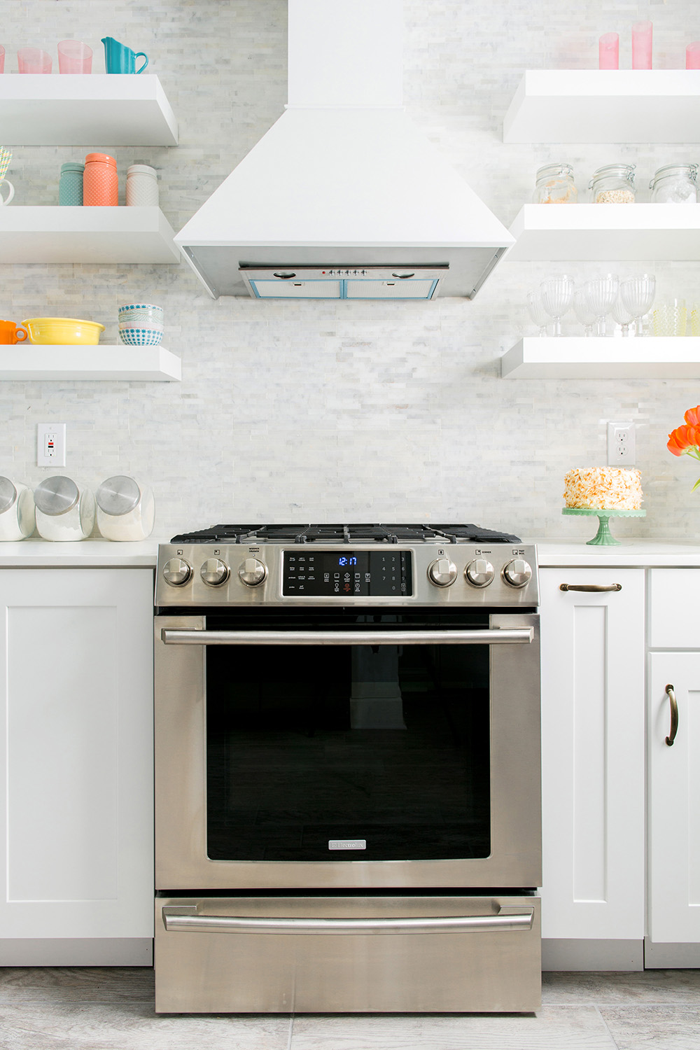 A silver kitchen oven with a white vent hood and white shelving on the wall.