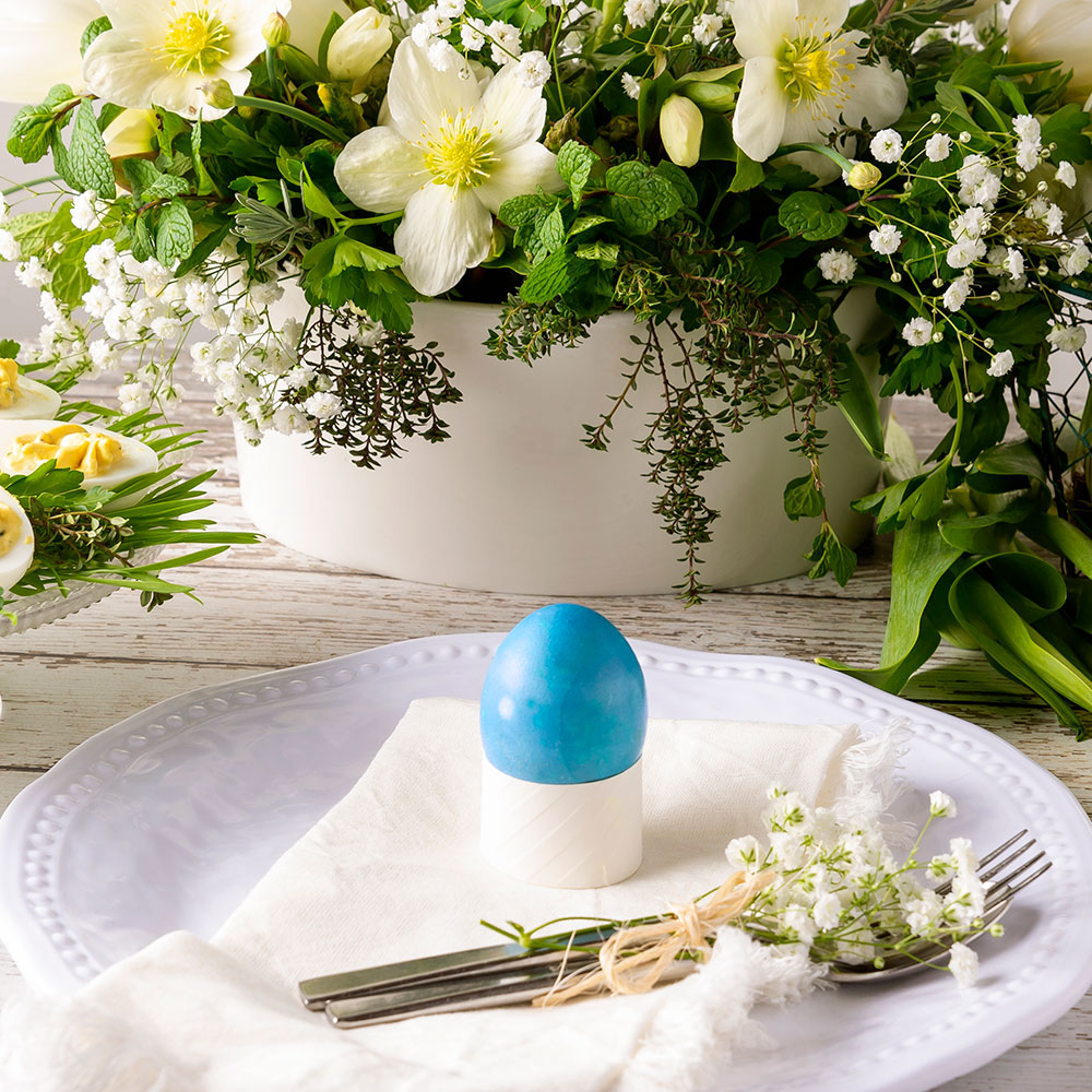 A blue Easter egg on a white plate with flowers in the background. A blue Easter egg on a white plate with flowers in the background.