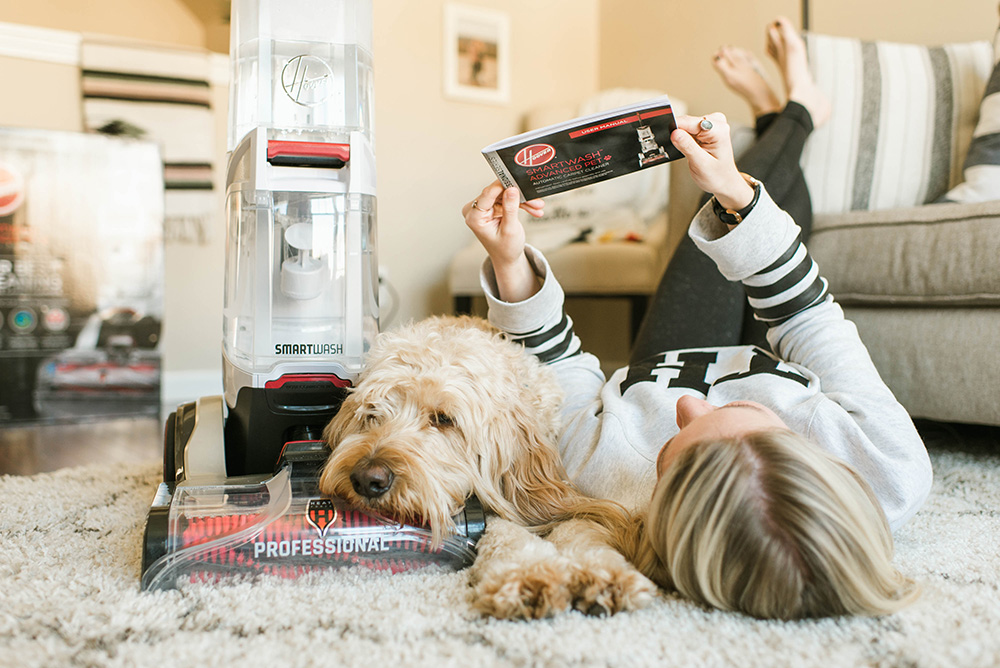 A woman and dog relaxing on a rug next to a carpet cleaner. 