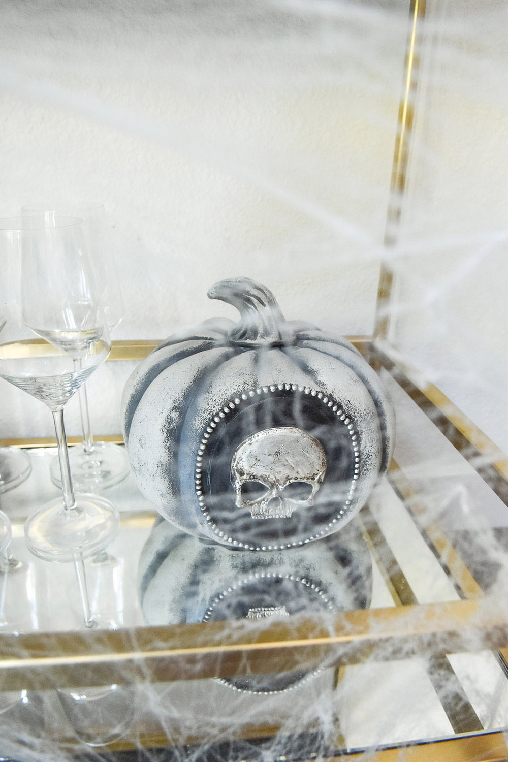 A Halloween pumpkin skull sits on the bottom of a glass and gold beverage cart.