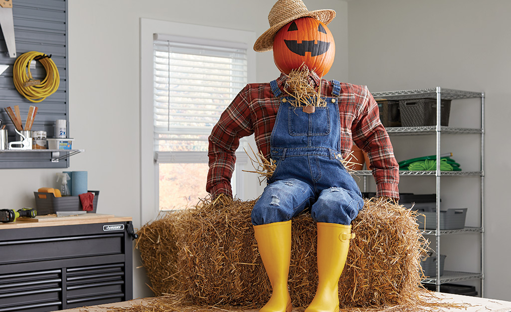 A scarecrow with a pumpkin head, overalls and yellow rain boots sits on a hay bale in a workshop.