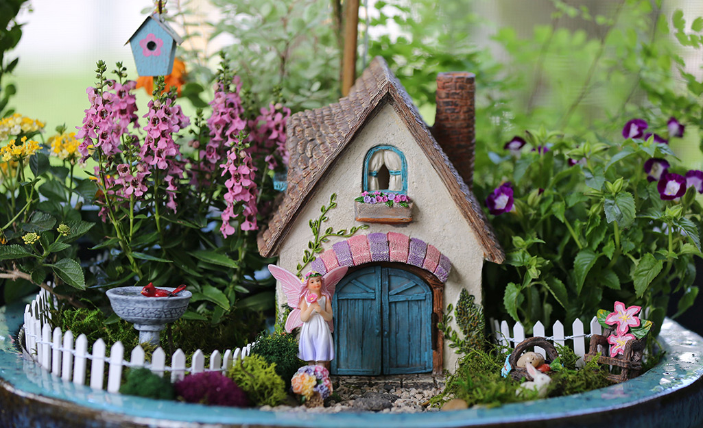A miniature garden includes a cottage, picket fence, bird bath and birdhouse along with flowering plants.