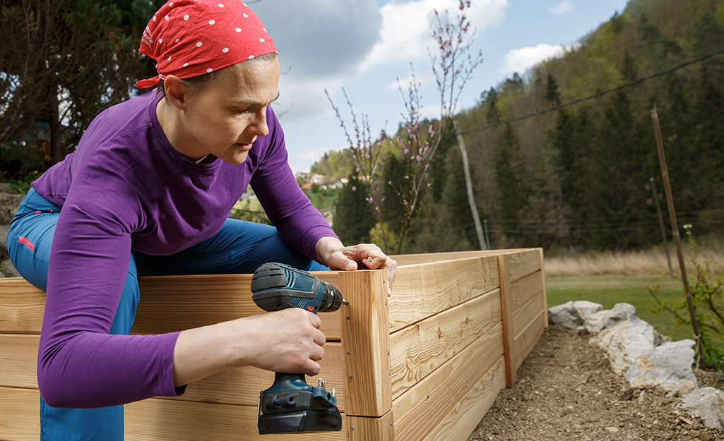 A woman uses a drill to connect the sides of a raised garden bed.