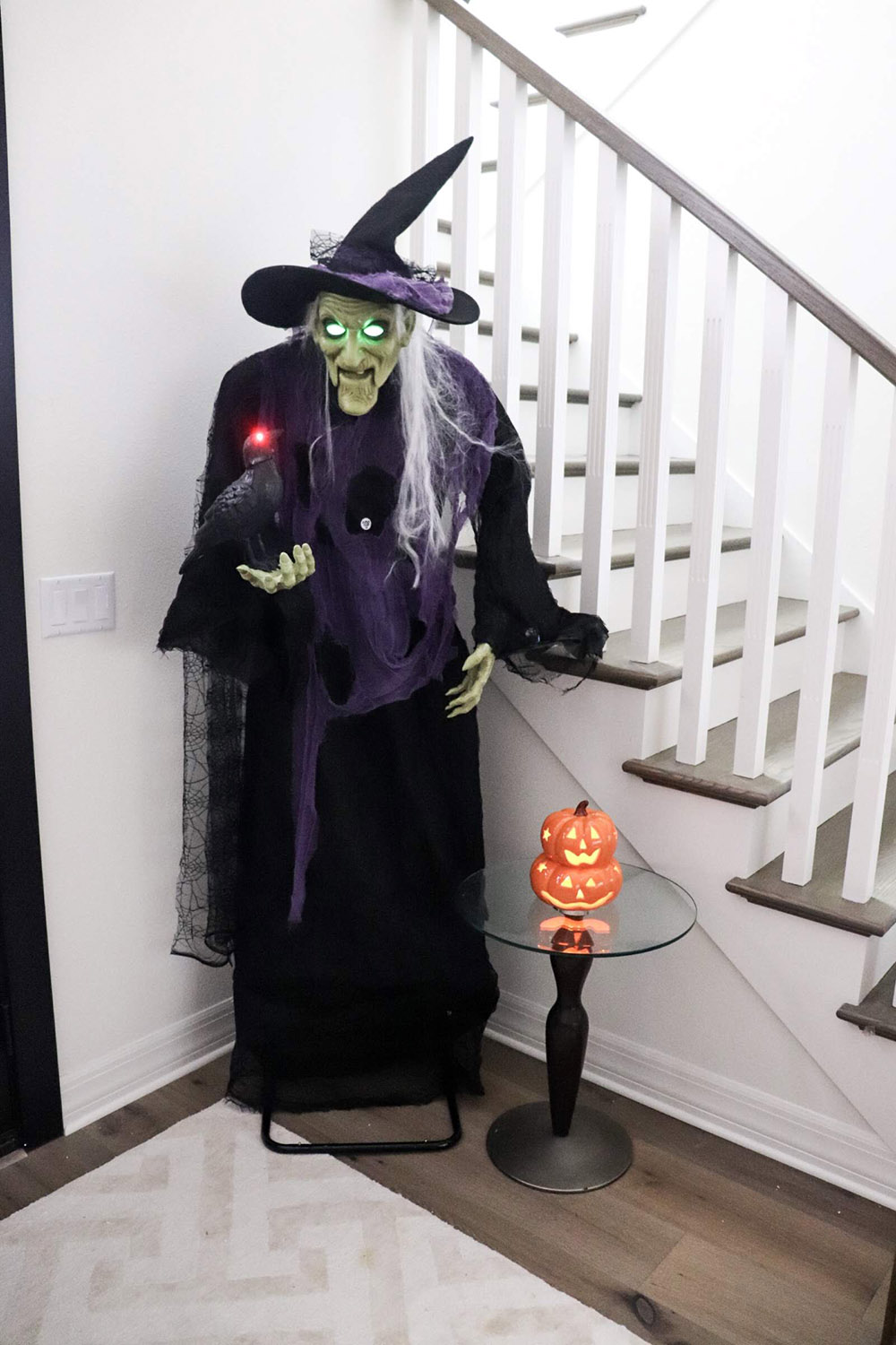 A tall witch with glowing eyes and a raven stand next to a staircase in the corner of a home.