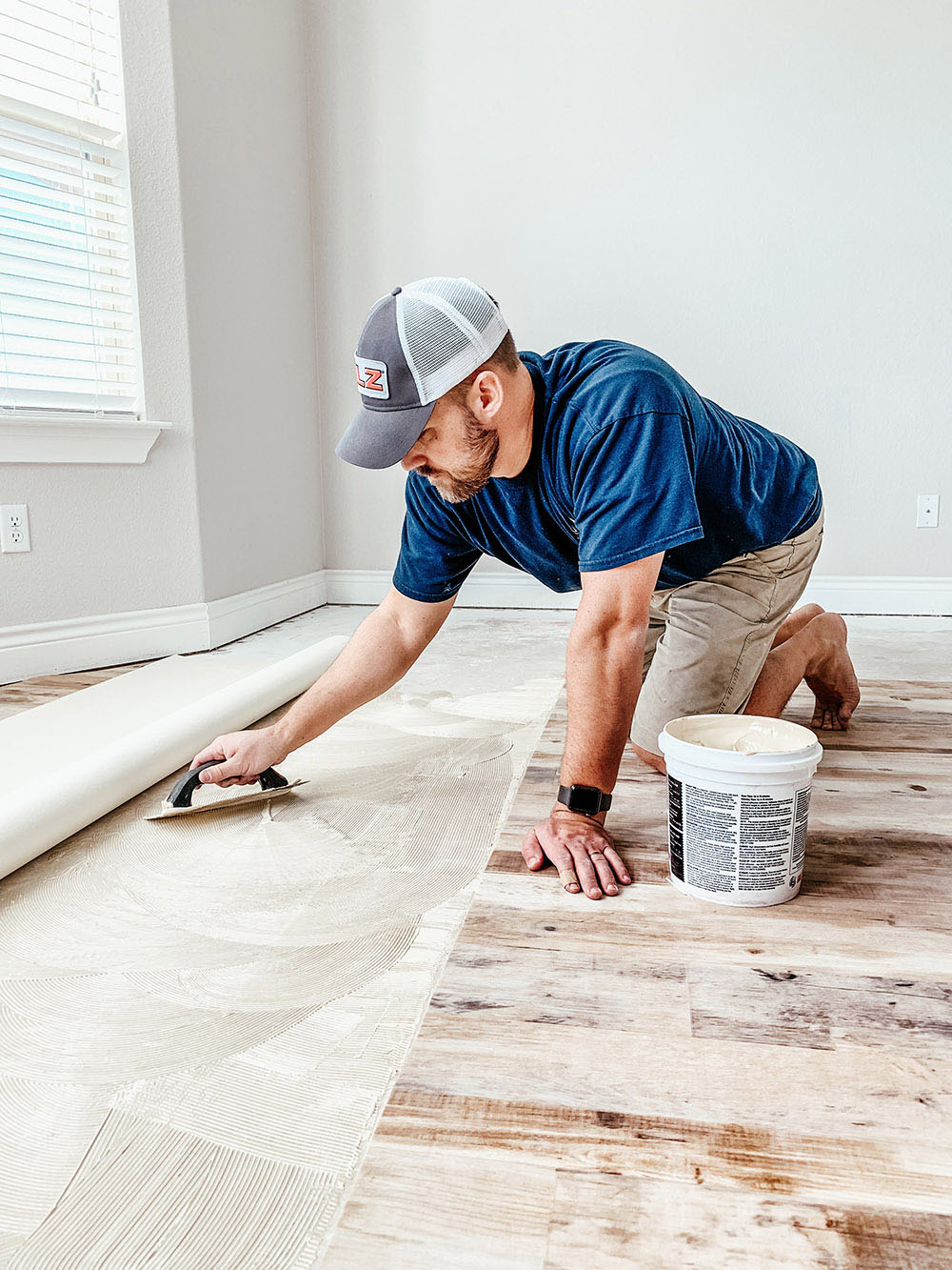 A man using a u-shaped trowel to apply adhesive to a concrete floor.