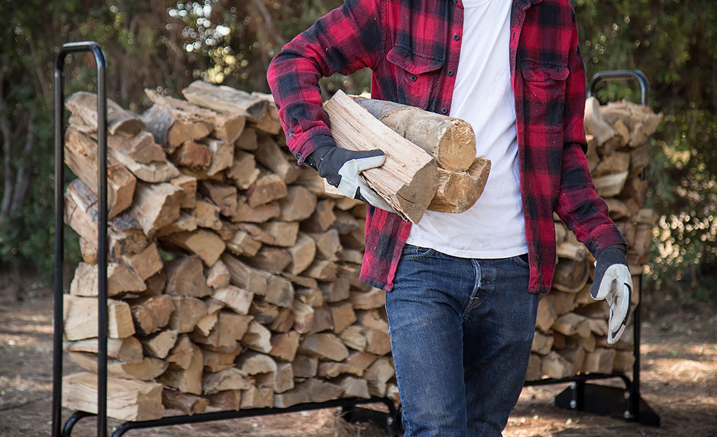 Someone carrying a few pieces of firewood away from a large rack of stacked, split firewood.