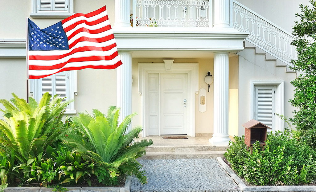 An American flag flies from a low flagpole standing among some leafy green plants beside a walkway leading to the front door of a house.