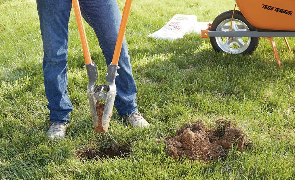 A man uses a post hole digger to make a hole in a yard for flagpole installation.