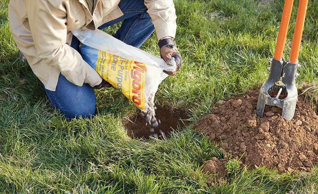 A man kneels down to pour landscaping rocks into a hole that was dug in a lawn for flagpole installation.