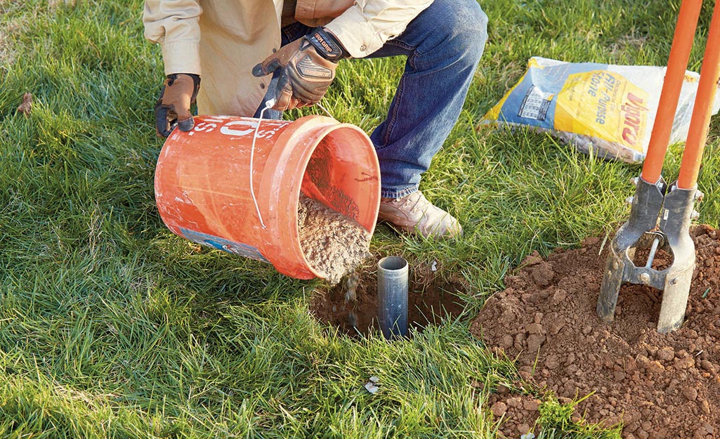 A man kneels in the grass as he pours concrete to set the ground sleeve during flagpole installation.