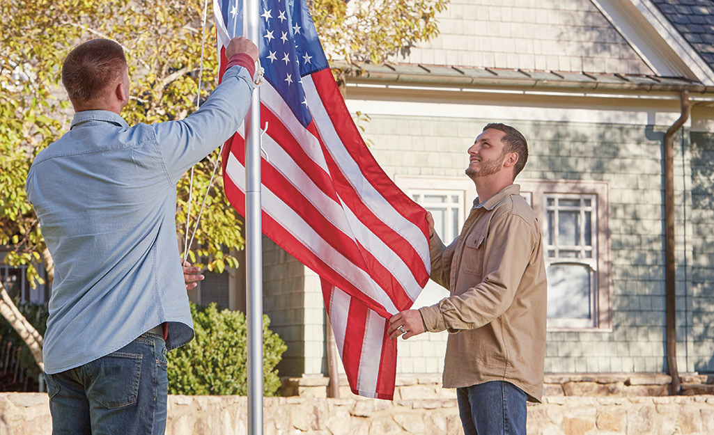 Two men raise an American flag on a flagpole installed in the front yard of a house. 