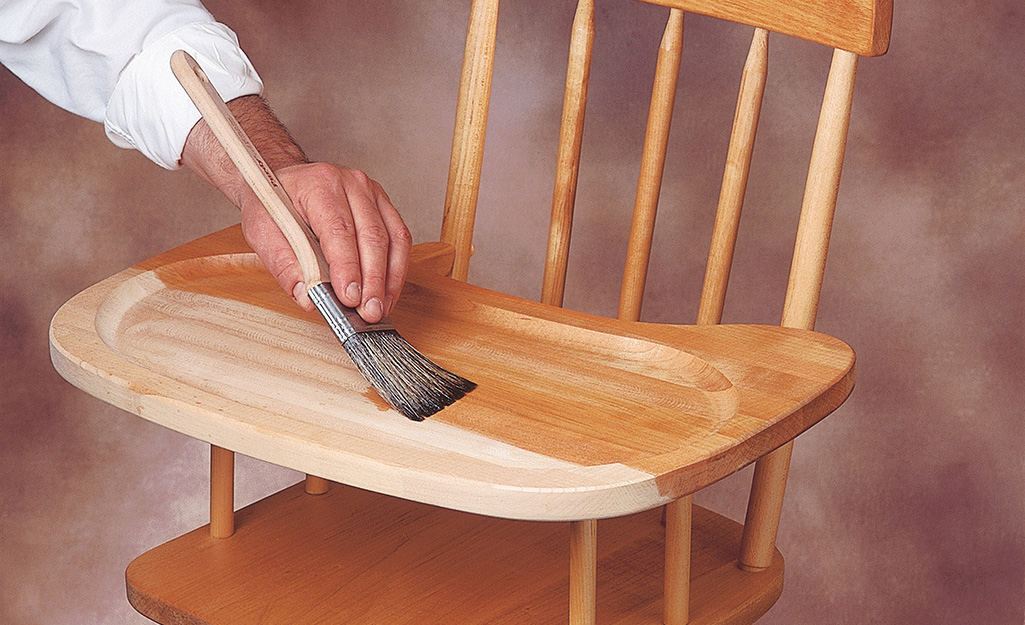 A person brushes finish onto a wooden high chair.