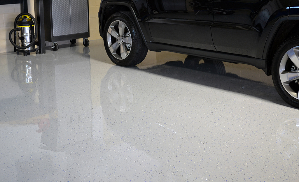 The shiny finish of a garage floor coated with gray epoxy paint shows the reflection of a black car.