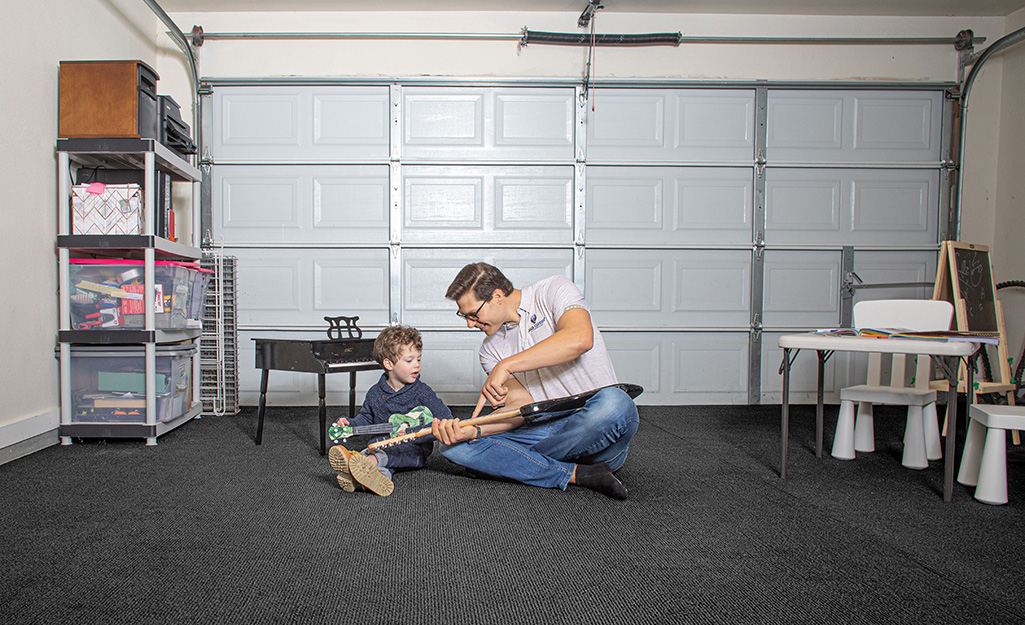 A man and a boy sit on the carpeted floor of a garage that has been converted into a playroom.