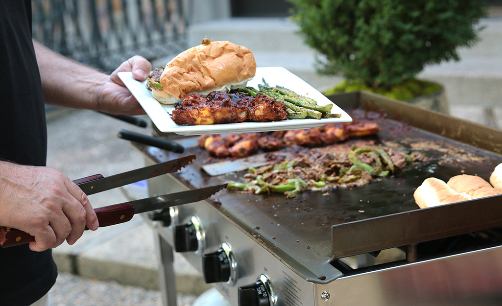 A person cooks meat and vegetables on a flat top gas grill.