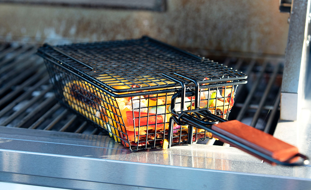 A long-handled grilling basket holds vegetables on a grill. 