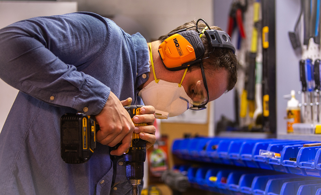 A person using a drill and wearing a mask and ear muffs for hearing protection.