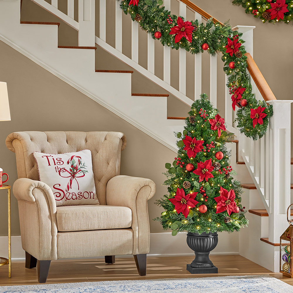 A potted Christmas tree with poinsettias by a staircase with garland and poinsettia blooms