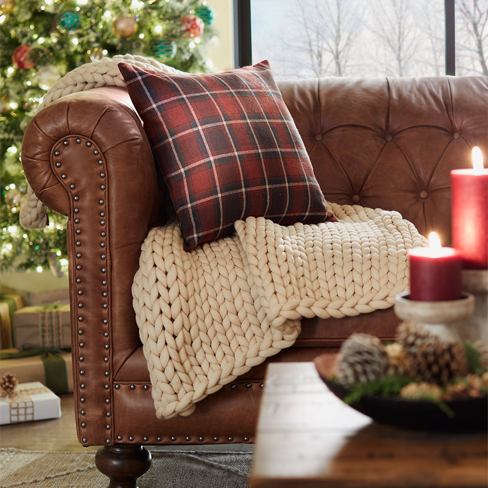 A plaid pillow sits on a white throw on a leather couch in front of a lit Christmas tree.