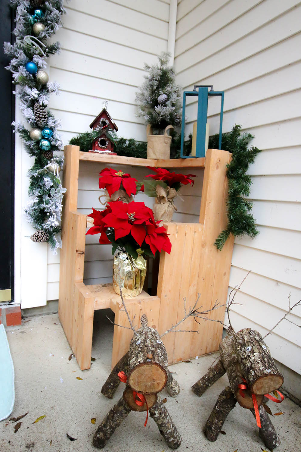 Two DIY wood slice reindeer standing in front of a three-tiered plant stand decorated with garland and poinsettias. Two DIY wood slice reindeer standing in front of a three-tiered plant stand decorated with garland and poinsettias.