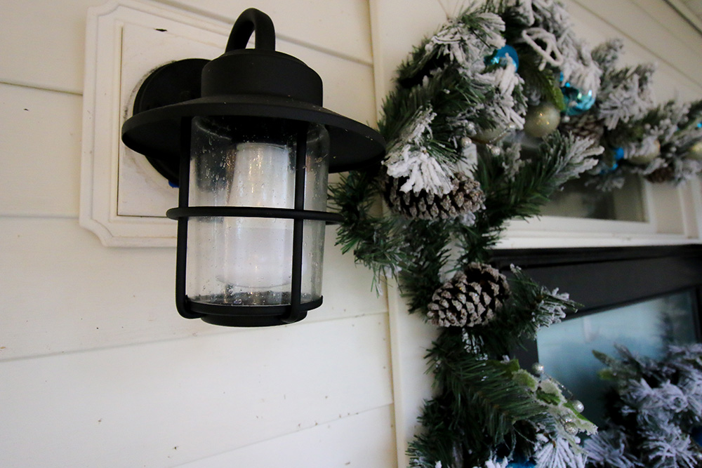 A coastal wall mounted lantern next to a front door surrounded by flocked garland. A coastal wall mounted lantern next to a front door surrounded by flocked garland.